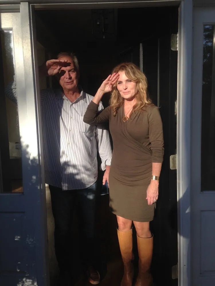 A person in a striped shirt and a person in a brown dress stand in a doorway, both raising hands in a salute.