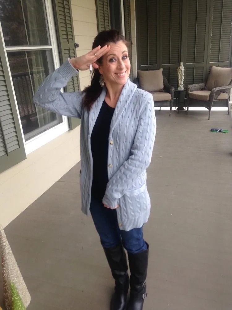 A person smiling and giving a salute while standing on a porch.