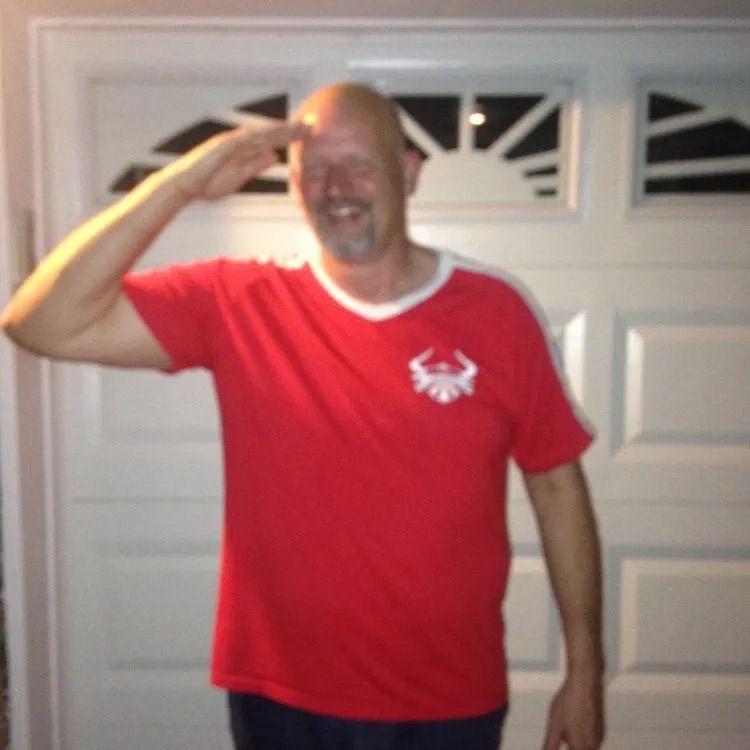 A person wearing a red t-shirt with a white logo salutes while standing in front of a white garage door.