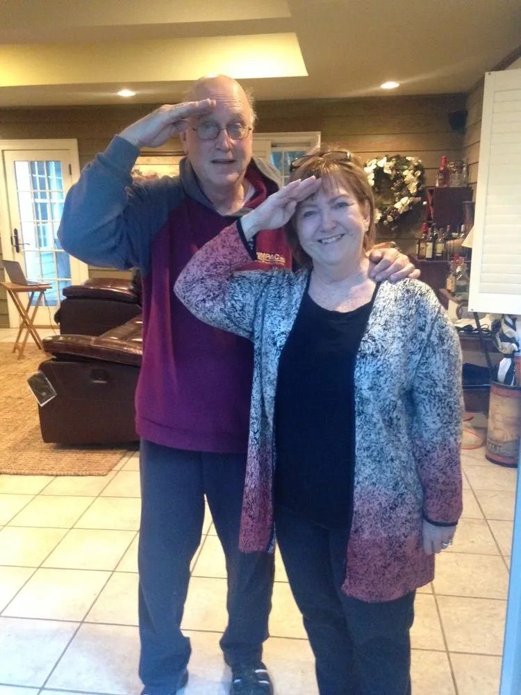 A man in a maroon hoodie and a woman in a patterned cardigan stand together indoors, both giving a hand salute and smiling.