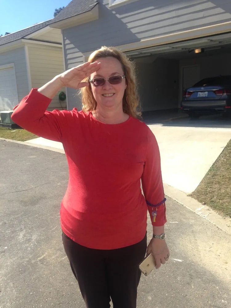 A person in a red long-sleeved shirt and sunglasses salutes outside a house with an open garage.