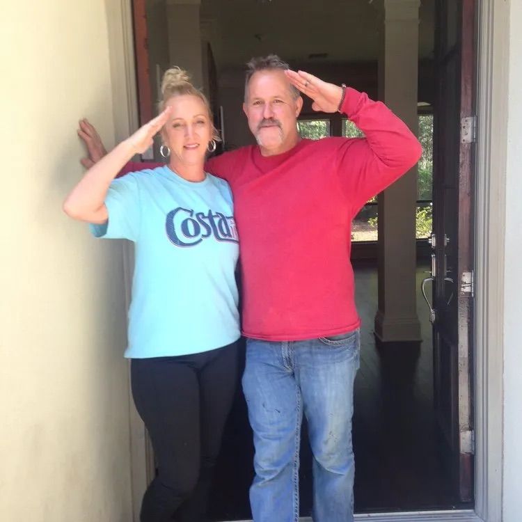 A woman and a man standing in a doorway saluting, both smiling, wearing casual clothes.
