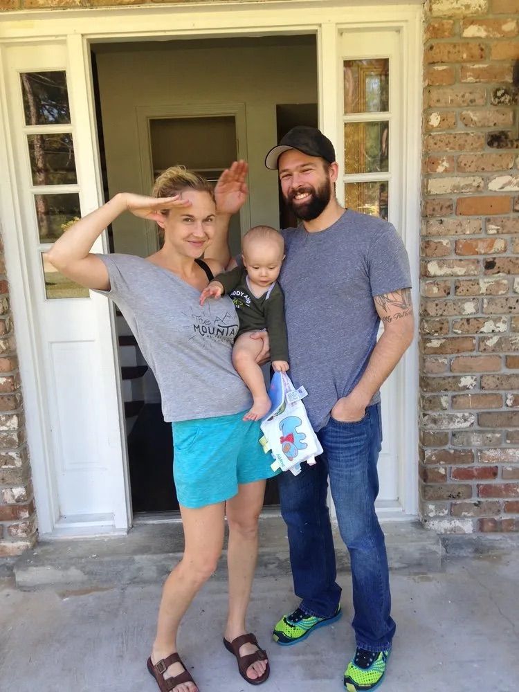 A family stands outside a doorway. A person salutes, another holds a baby, and a bearded person stands next to them.