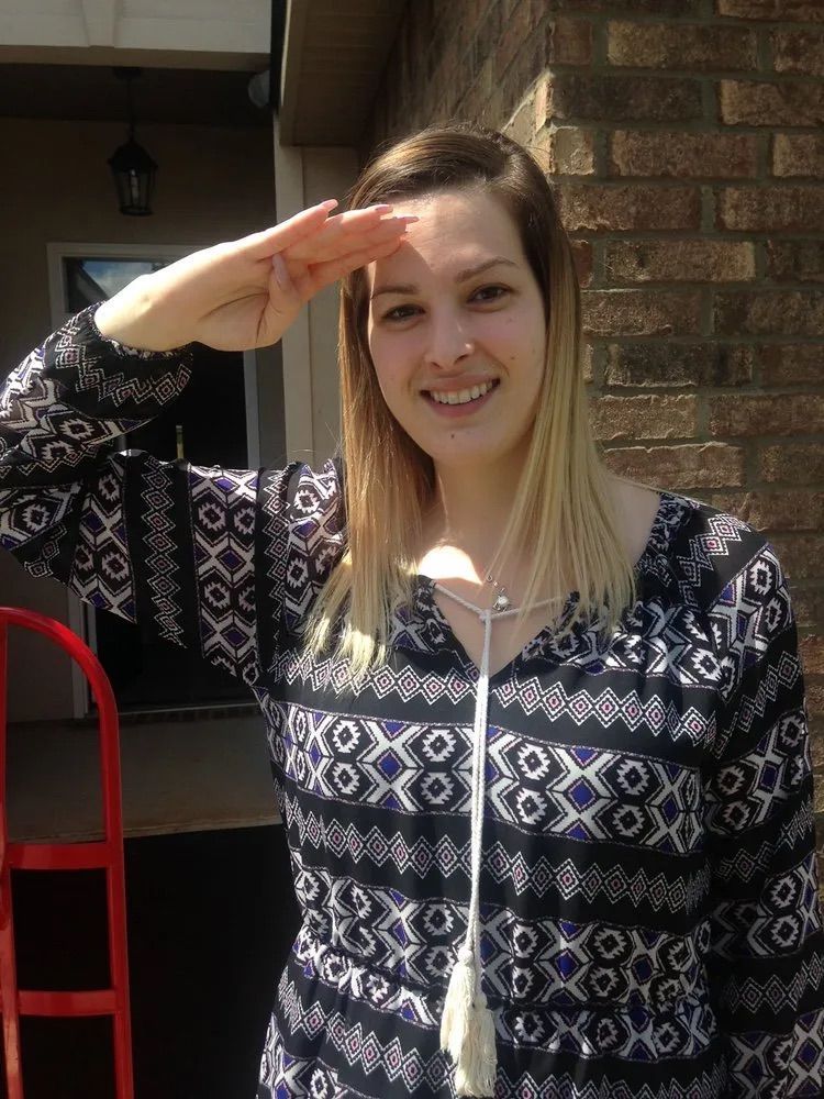 A person with blonde hair wearing a patterned blue and white top saluting in front of a brick building.