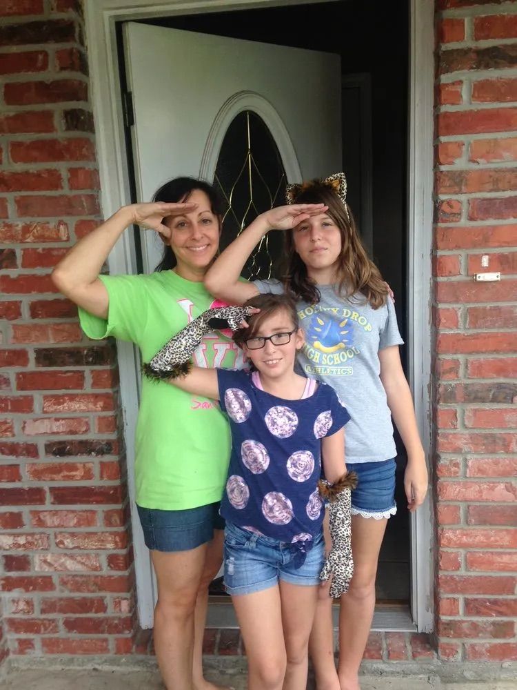 Three people stand in front of a brick building, posing with hands saluting, wearing casual shirts, shorts, and accessories.