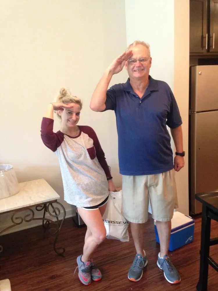 A person in a maroon and white shirt and a person in a blue polo shirt stand side-by-side, both saluting in a room.