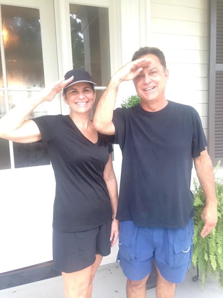 A smiling couple standing outdoors and saluting toward the camera while wearing casual clothing.