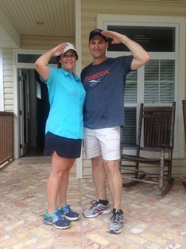 Two people stand on a porch saluting, wearing casual athletic clothes and baseball caps.