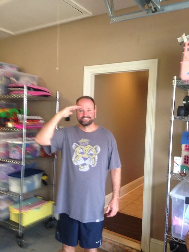 A smiling person wearing a grey tiger-graphic t-shirt and blue shorts salutes while standing in a garage.