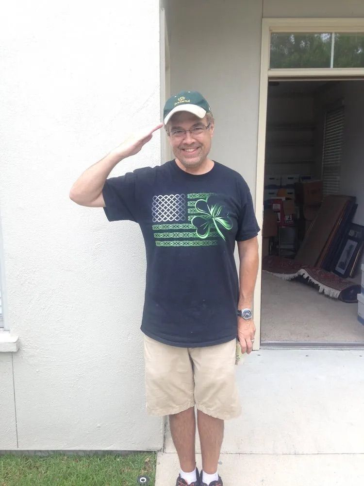 A person wearing a green hat and a dark green-themed American flag shirt salutes while standing outside a doorway.