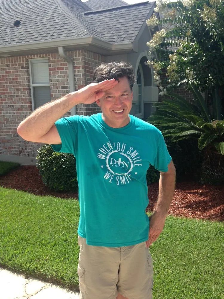 A smiling person wearing a teal t-shirt with text, standing in front of a house, saluting with their right hand.
