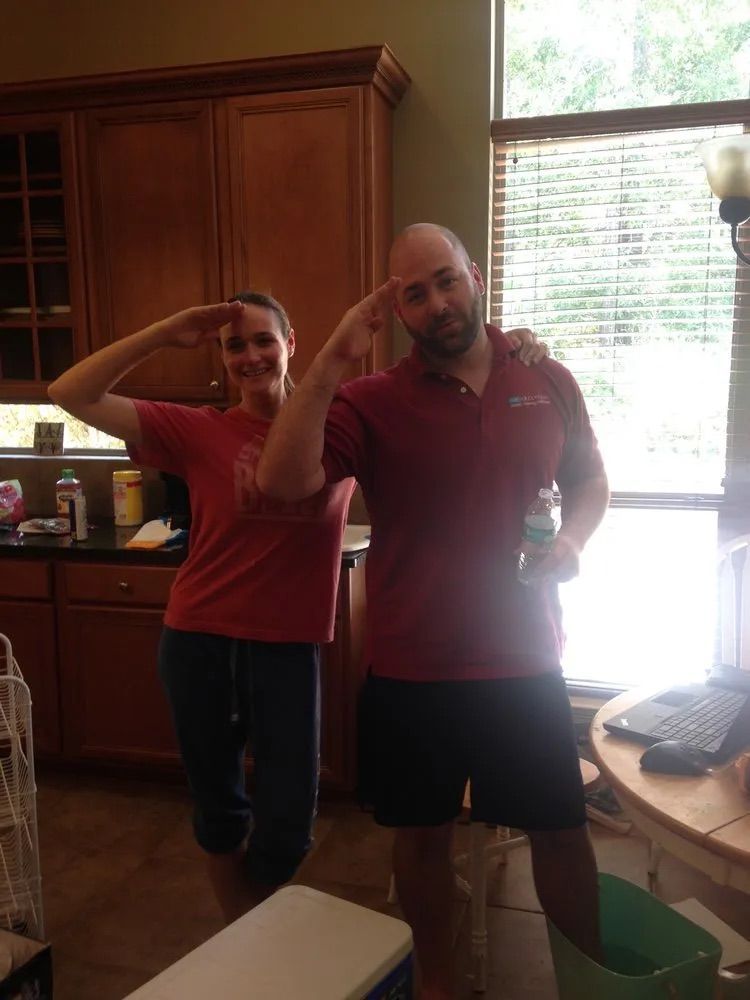 Two people in red shirts stand in a kitchen, both giving a hand salute while looking toward the camera.