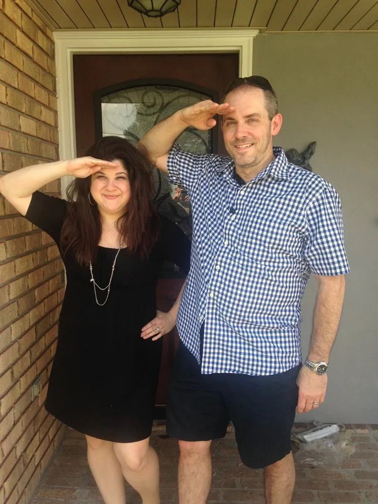 A person in a black dress and another in a blue checkered shirt standing on a porch, both saluting with their hands.