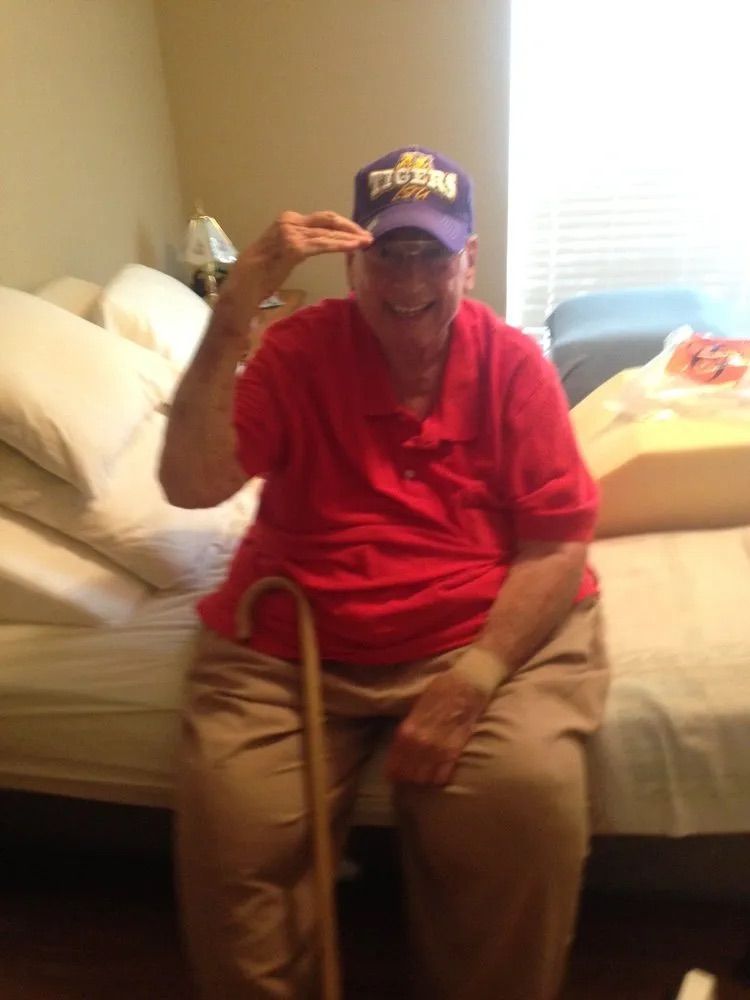 An individual in a red polo shirt and purple cap salutes while sitting on a bed with a wooden cane nearby.