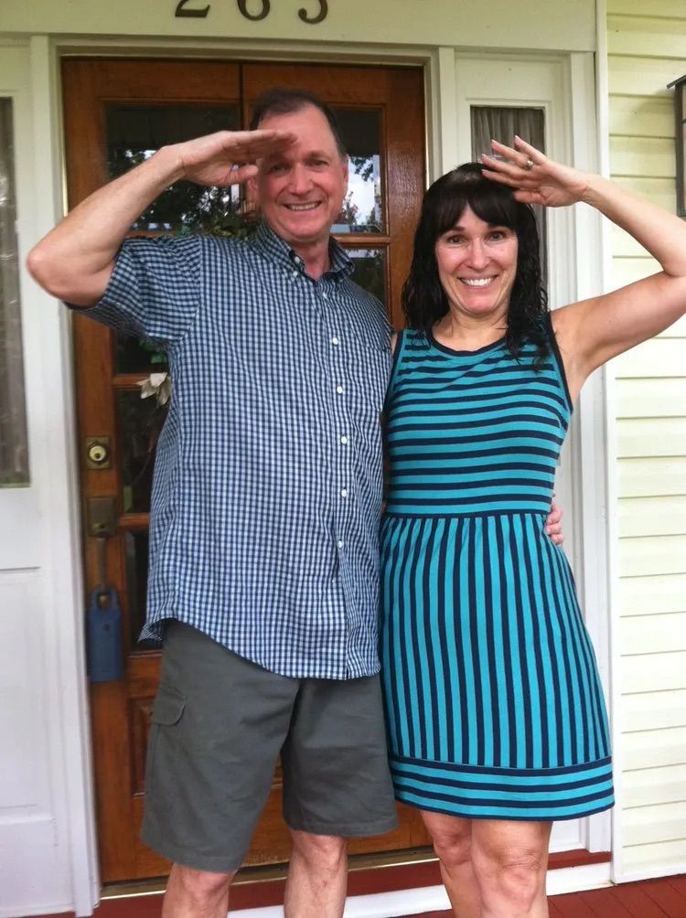 A person in a plaid shirt and shorts and a person in a blue striped dress standing by a door, both saluting the camera.