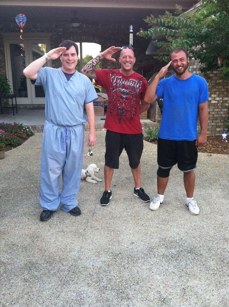 Three people standing outdoors on a patio, smiling and saluting while a small white dog rests on the ground behind them.