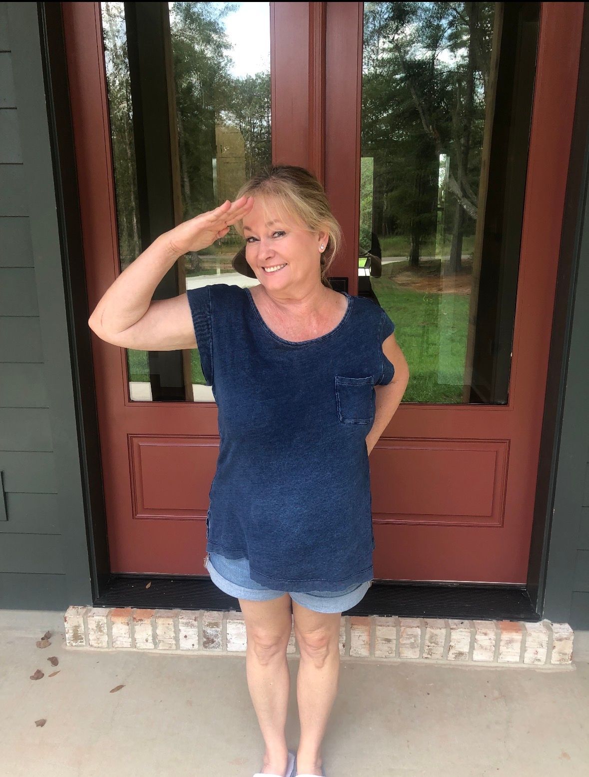 A person stands in front of a red door, saluting with one hand while smiling.