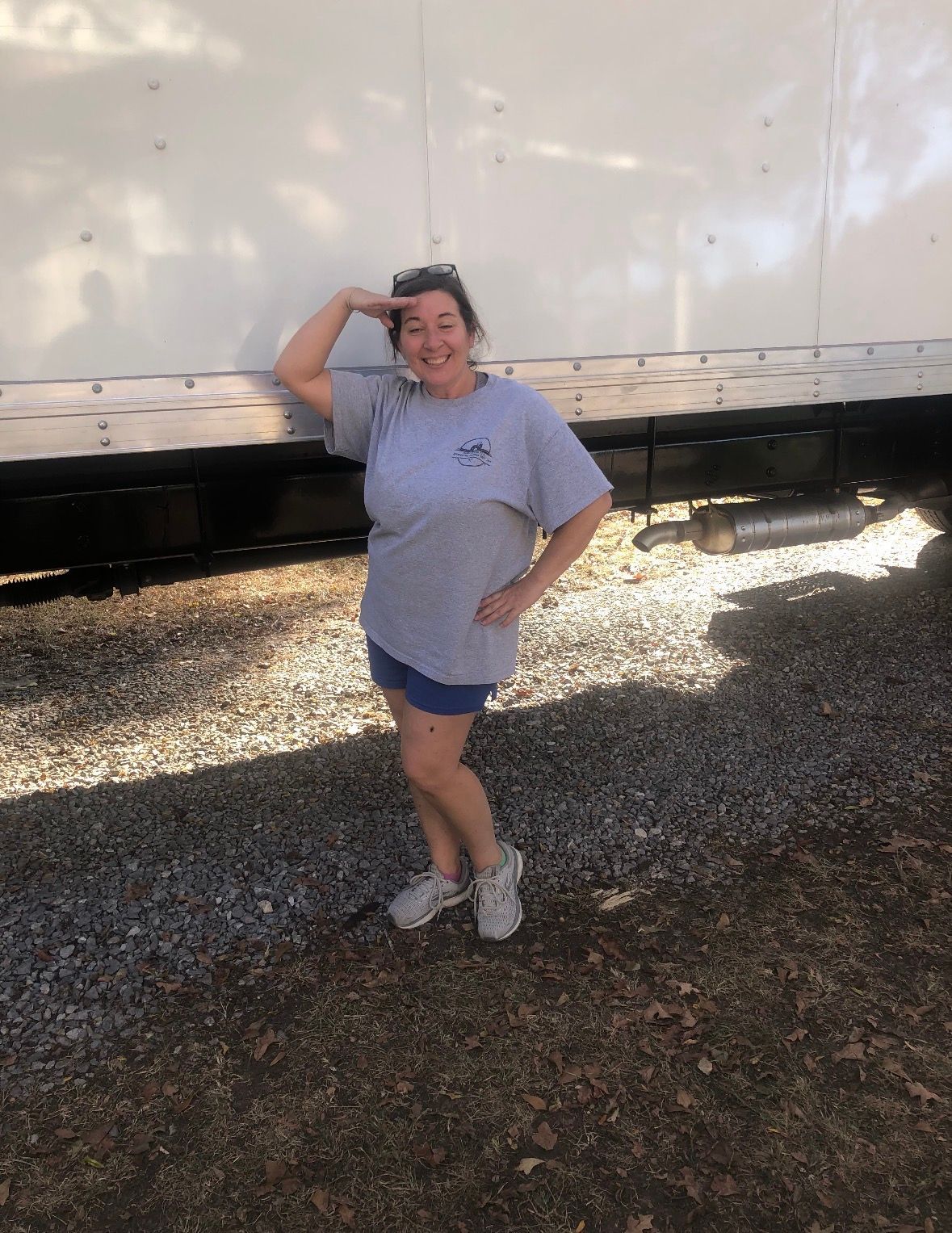 A smiling person wearing a grey t-shirt and blue shorts salutes while standing on gravel in front of a white truck trailer.