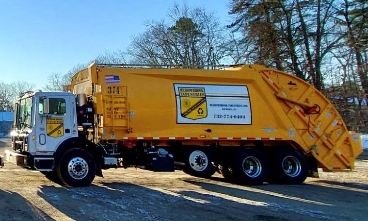 A yellow side-loader garbage truck parked on a dirt lot, featuring a company logo and a phone number on its side.