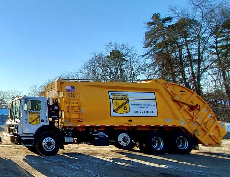 A yellow sanitation truck parked on a gravel lot under a clear blue sky, with trees in the background.