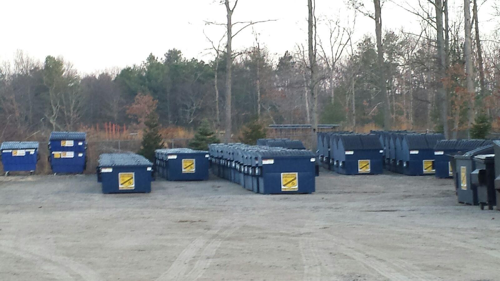 Many blue dumpsters lined up in rows on a large, gravel outdoor storage lot near trees.