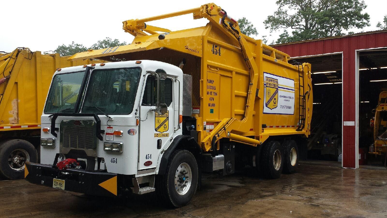 A yellow side-loader garbage truck with a white cab is parked outdoors in front of a red building.