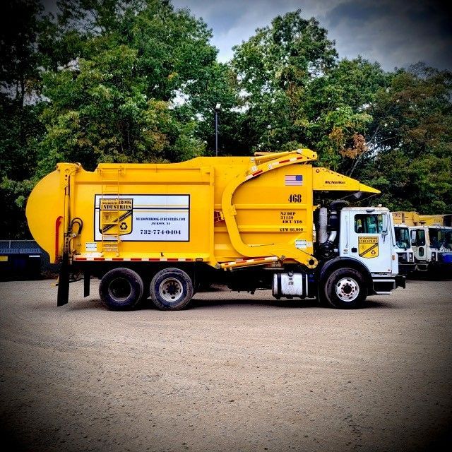 A bright yellow residential garbage truck parked on a gravel lot with trees in the background.