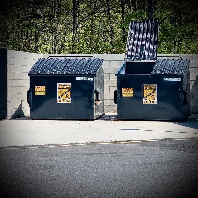 Two dark blue commercial dumpsters sit in a concrete enclosure against a wall, with one lid open.