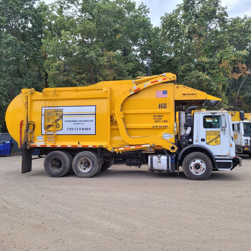 A yellow residential side-loader garbage truck parked on a gravel lot in front of a tree line.