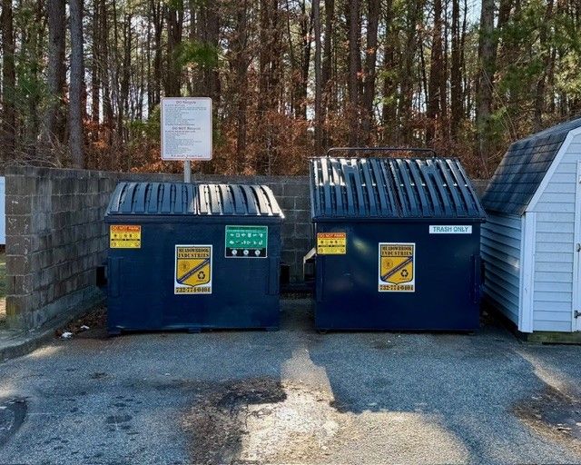 Two blue industrial dumpsters placed on an asphalt surface in front of a brick wall and a wooded area.