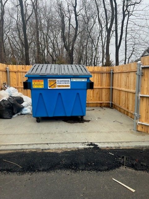 A blue dumpster sits on a concrete pad enclosed by a wooden fence, with some trash bags on the left.