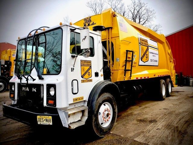 A white Mack garbage truck with a large yellow compactor body parked on a dirt lot.