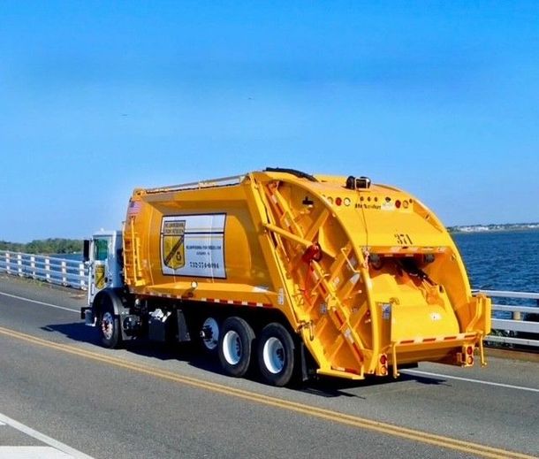 A yellow garbage truck driving across a bridge over a body of water under a clear blue sky.