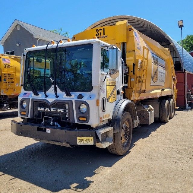 A white and yellow Mack garbage truck parked on a gravel lot under a clear sky next to a warehouse building.