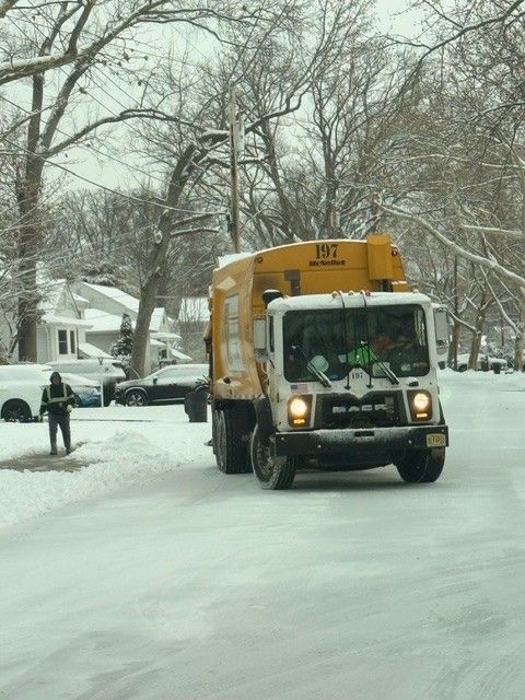 A yellow garbage truck drives down a snow-covered suburban street as a worker walks along the side of the road.