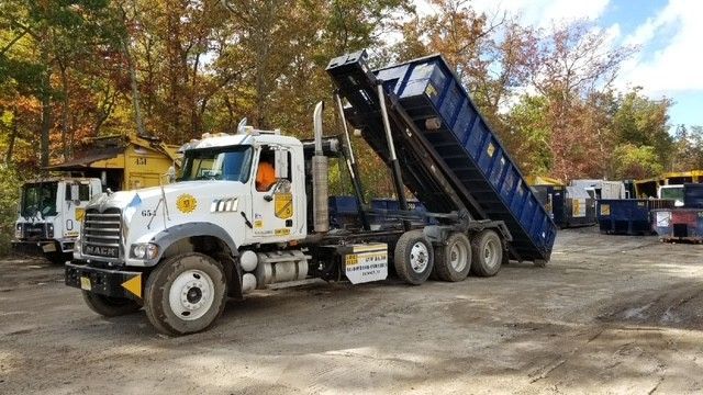 A white Mack roll-off truck with a tilted blue dumpster container in an outdoor yard with trees in the background.