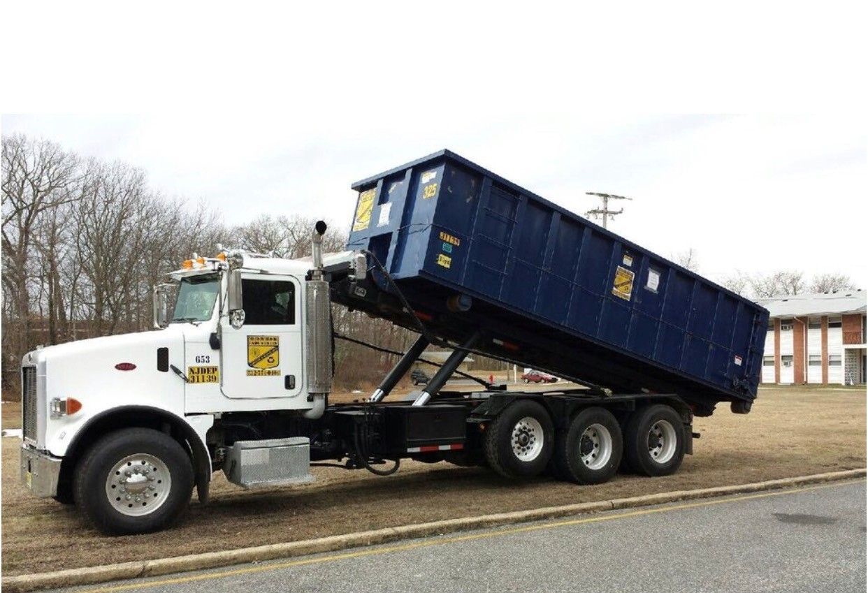 A white roll-off truck with a blue dumpster elevated at an angle, parked on a gravel lot near a building.