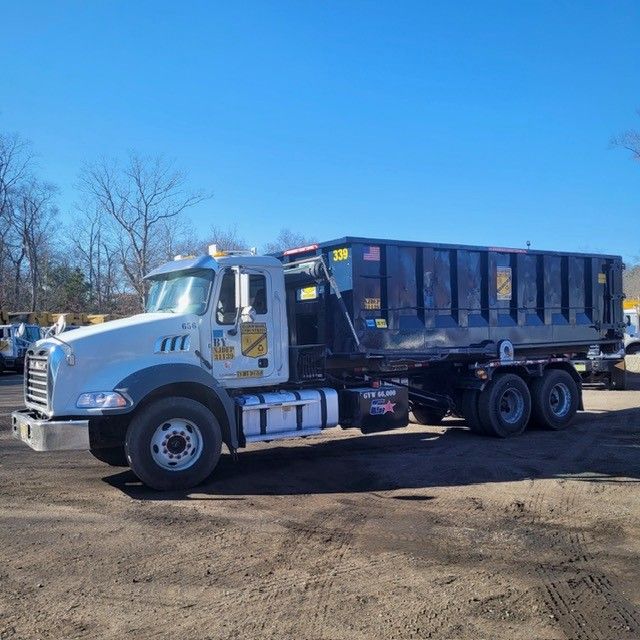 A white roll-off dump truck with a large black container parked on a dirt lot under a clear blue sky.