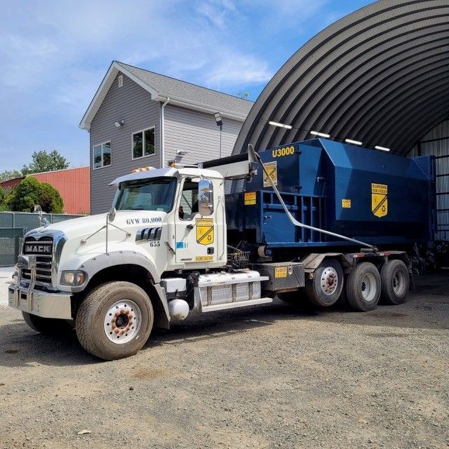 A white Mack truck with a large blue dumpster container parked on a gravel lot next to a warehouse.