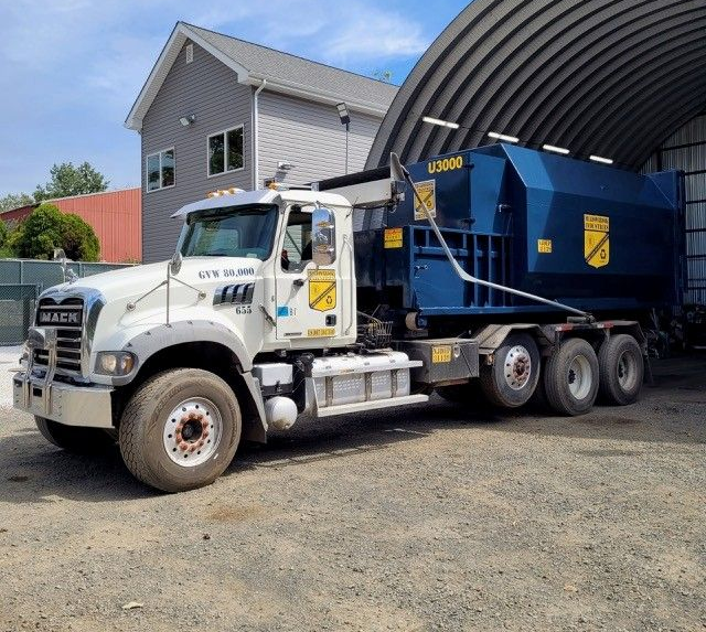 A white Mack truck with a large blue dumpster unit parked on a gravel lot next to a metal building.