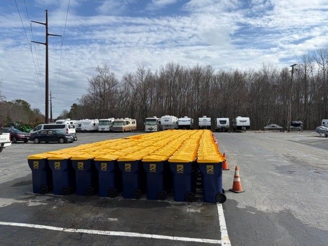 Rows of dark blue trash bins with yellow lids are lined up in an outdoor parking lot with RVs in the background.