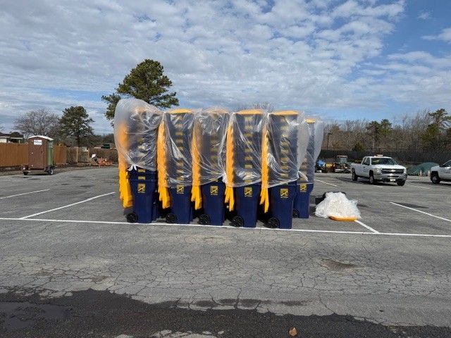 A row of new blue and yellow trash bins wrapped in plastic stands in a parking lot under a cloudy sky.