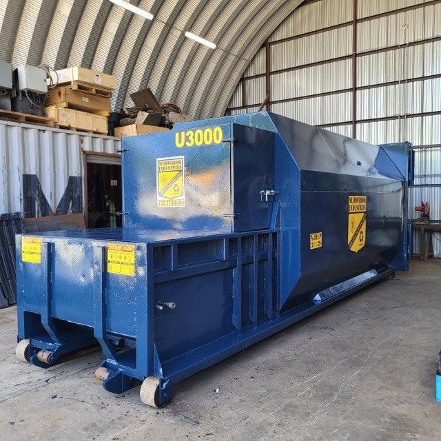 A blue industrial trash compactor, labeled U3000, sits on a concrete floor inside a large warehouse storage building.