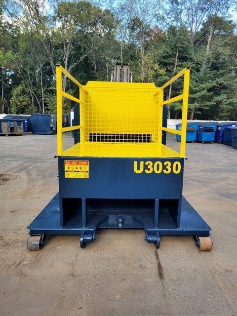 A yellow and blue steel forklift work platform with railings sits on concrete with trees in the background.