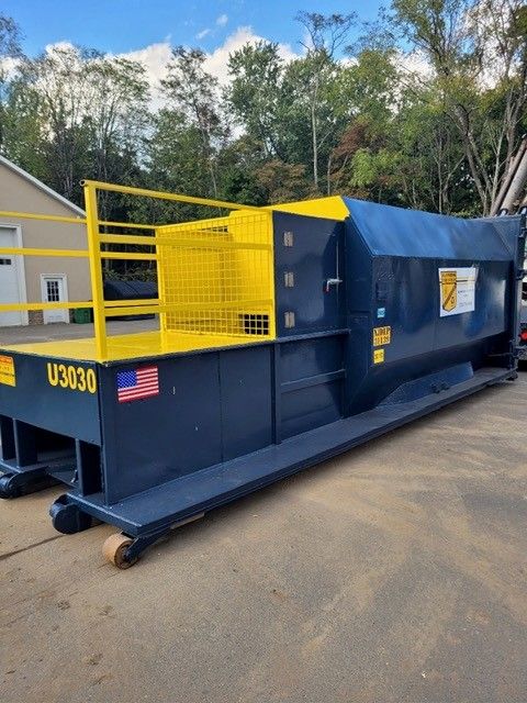 A dark blue industrial waste compactor with yellow safety railings outdoors in a paved lot.