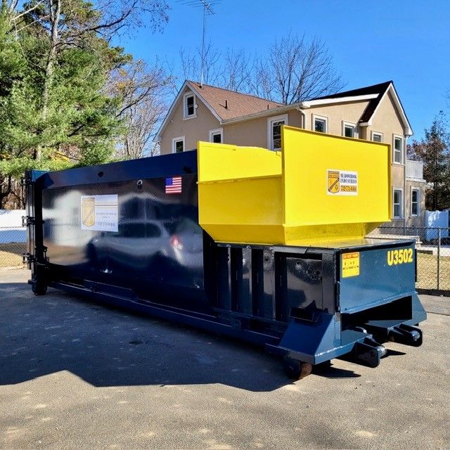 A large dark blue industrial trash compactor with a bright yellow hopper, parked in a driveway before a house.