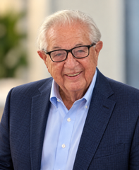 A person wearing glasses, a white collared shirt, a red tie, and a dark red jacket, smiling against a light background.