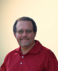 A smiling person with glasses and a goatee, wearing a red polo shirt against a plain, light background.