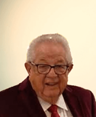 A person wearing glasses, a white collared shirt, a red tie, and a dark red jacket, smiling against a light background.