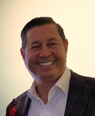A person with short, dark hair smiling, wearing a light-colored collared shirt and a dark textured blazer.